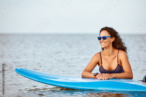 Young smiling woman in sunglasses with surfboard standing on the beach