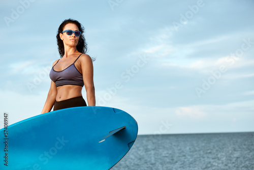 Young smiling woman in sunglasses with surfboard standing on the beach © Georgii