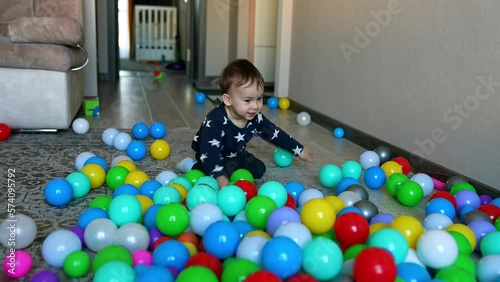 Funny adorable toddler comes up to the multiple balls scattered on the floor. Kid sits down on his knees and kicking the balls. photo