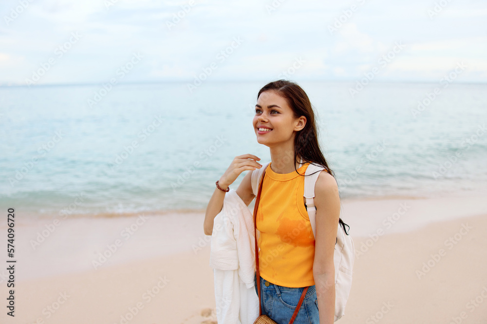 Woman smile with teeth after swimming in the ocean with a backpack in a wet yellow tank top and denim shorts walks along the beach, summer vacation on an island by the ocean in Bali sunset Woman smile with teeth after swimming in the ocean with a backpack in a wet yellow tank top and denim shorts walks along the beach, summer vacation on an island by the ocean in Bali sunset