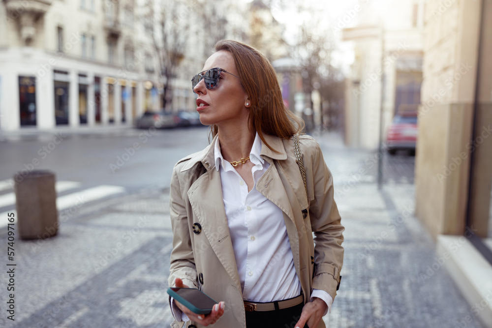 Fototapeta premium Fashionable woman in sunglasses is standing with phone on city street background and looking at side