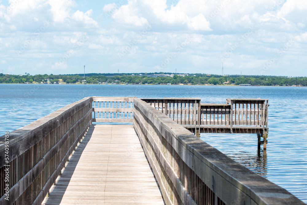 A wooden plank wharf or pier with wood rails jutted into the still blue ocean water.  The blue sky and clouds are reflected in the ocean water. There's a land with trees in the background.  