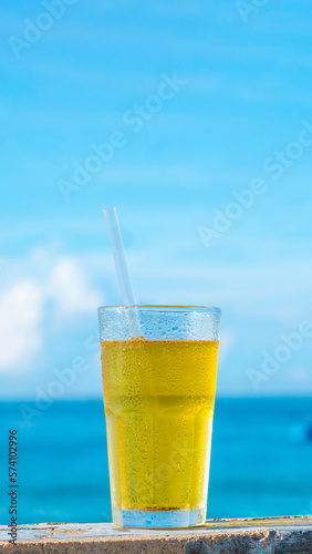 Big glass drink plastic straw green tea looks like yellow beer or kombucha blue sea white clouds background water drops condensed