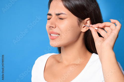 Young woman cleaning ear with cotton swab on light blue background, closeup