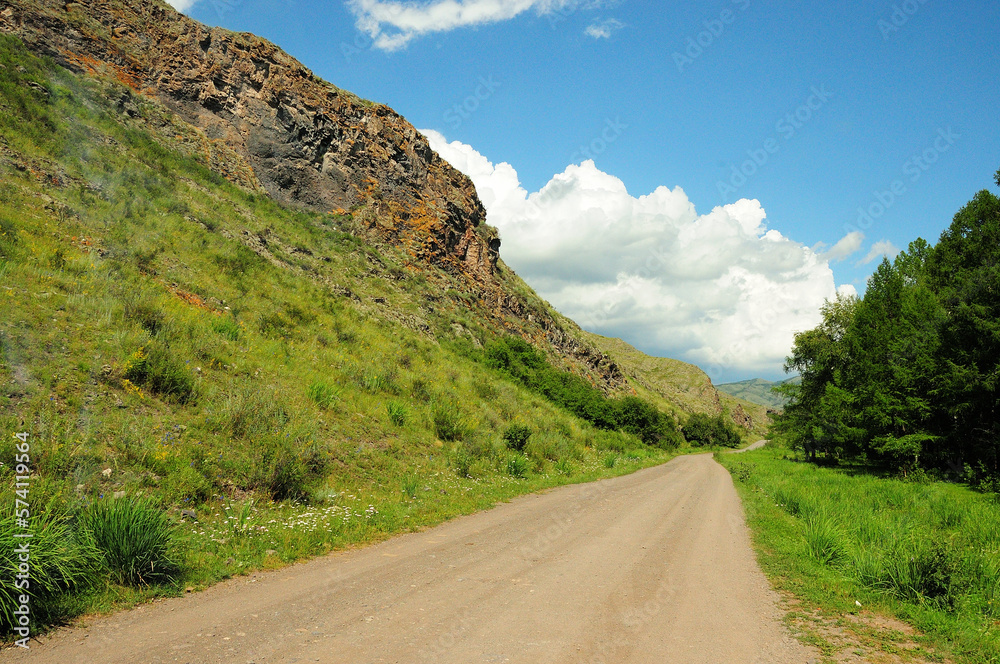 Narrow gravel road going through the edge of the forest at the foot of a high hill with rocky formations on a warm summer day.