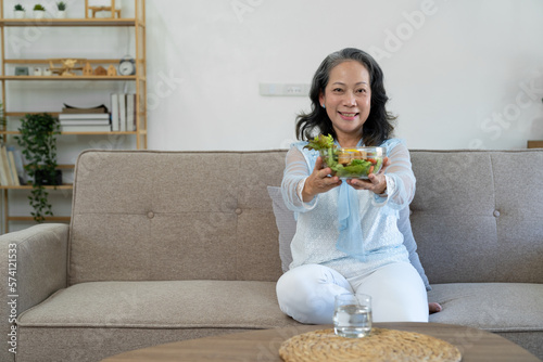 Smiling Asian elderly woman happily eating fruit and vegetable salad Charming to sit on the sofa and relax at home while eating with a glass of water. relaxation concept home interior.