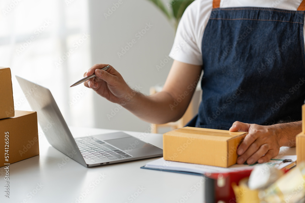 Entrepreneur, small business, SME, young freelance Asian man working from home carrying parcels for delivery taking note and checking customer list from laptop for verification online parcel delivery
