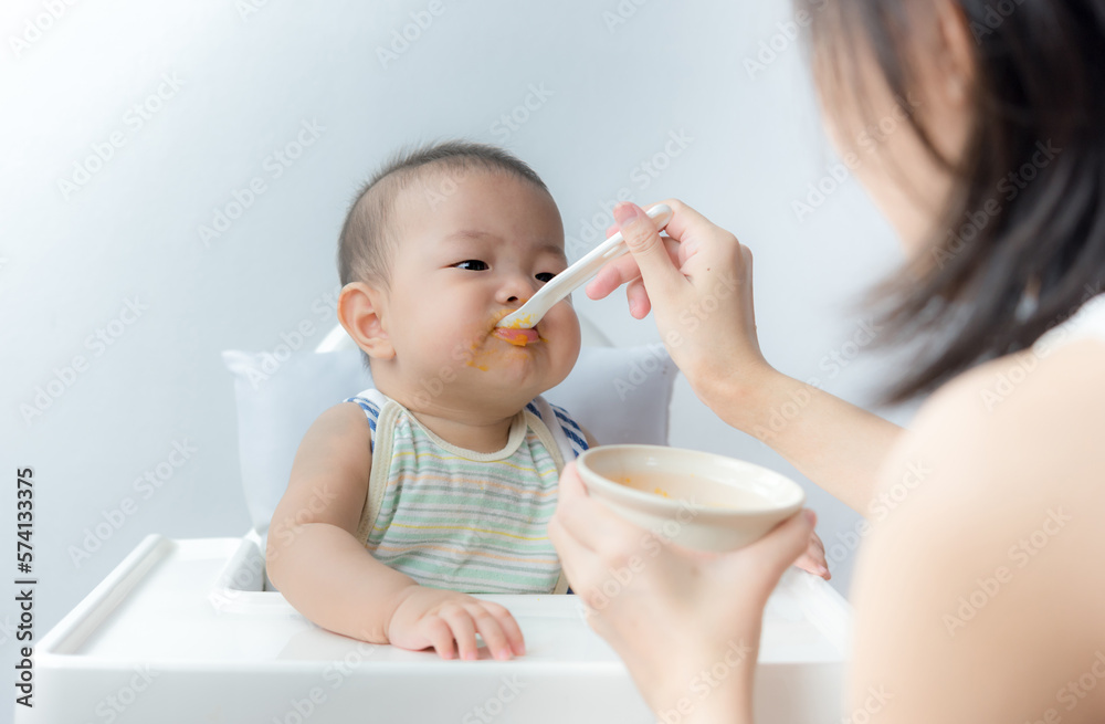 Mother feeding baby with spoon, baby boy child sitting eating food on chair at home Mother feeding baby with spoon, baby boy child sitting eating food on chair at home