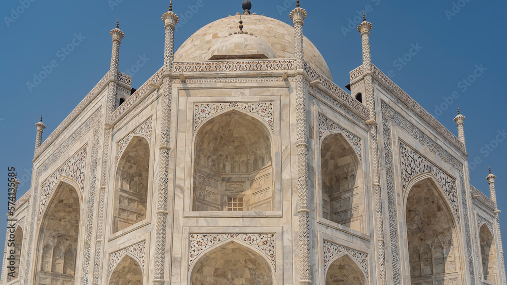 The ancient white marble Taj Mahal against the blue sky. Symmetrical arches, domes and spires are visible. On the walls of the mausoleum there are ornaments and inlays of precious stones. India. Agra 