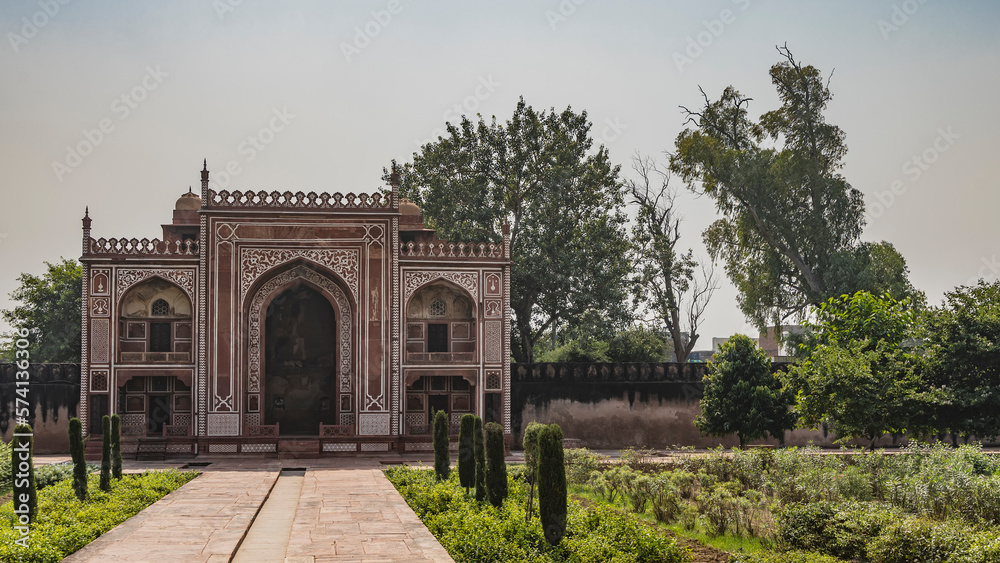 The ancient building in the Itmad-Ud-Daula tomb complex is made of red sandstone decorated with white ornaments. Arches, domes, latticed windows are visible. Green vegetation. India. Agra