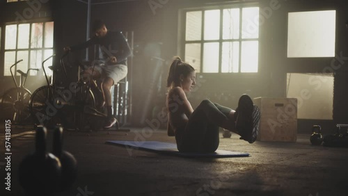 Woman wearing sportswear doing kettelbell twist on yoga mat at gym in slow-motion while two more men exercising in the background photo