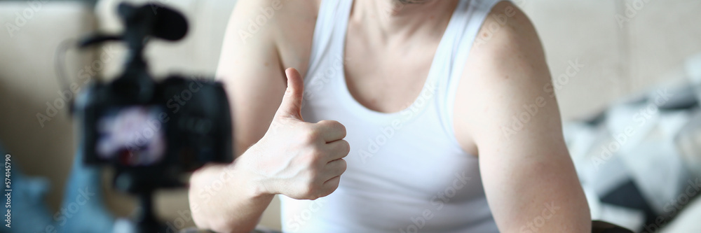 Athlete man holds thumbs up and raises dumbbells sitting on ball at home in front of camera