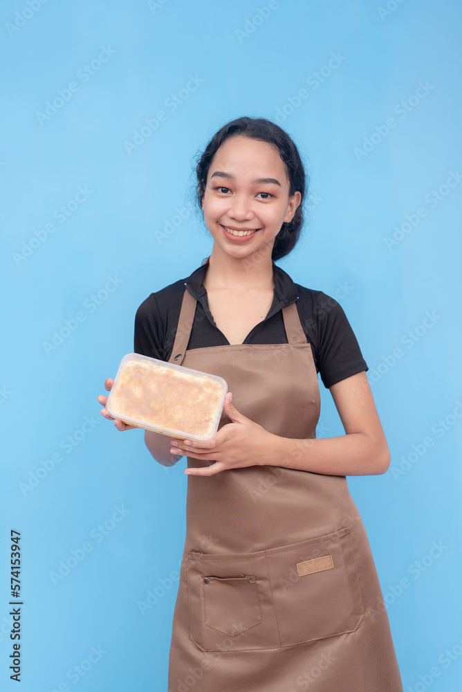 A young and friendly asian female baker or waitress presenting a tub of mango grahams. Wearing a brown apron and black shirt and against a light blue background. A young and friendly asian female baker or waitress presenting a tub of mango grahams. Wearing a brown apron and black shirt and against a light blue background.