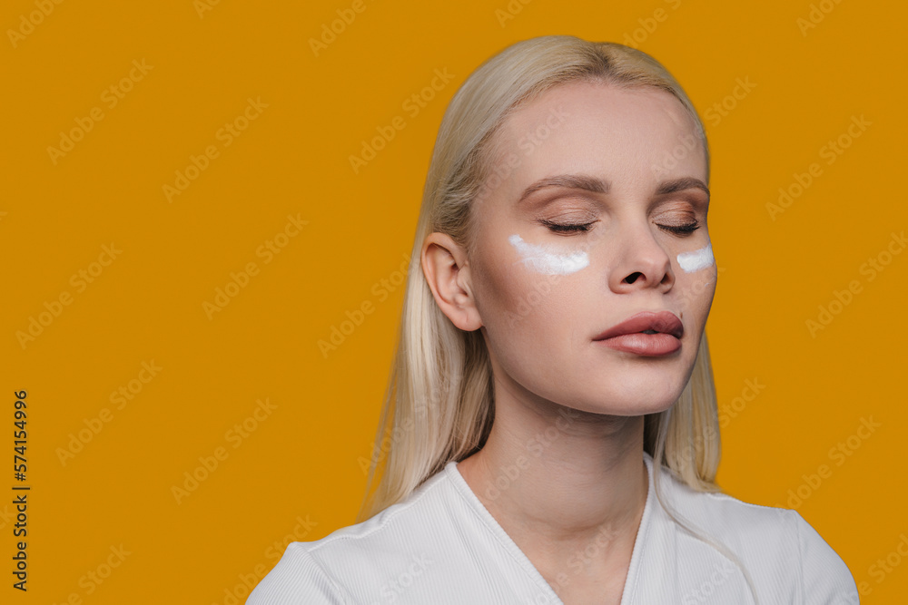 Caucasian blonde woman with cream on her cheek, enjoying skin care, posing with closed eyes isolated over yellow background. Beauty, tenderness concept. Caucasian blonde woman with cream on her cheek, enjoying skin care, posing with closed eyes isolated over yellow background. Beauty, tenderness concept.
