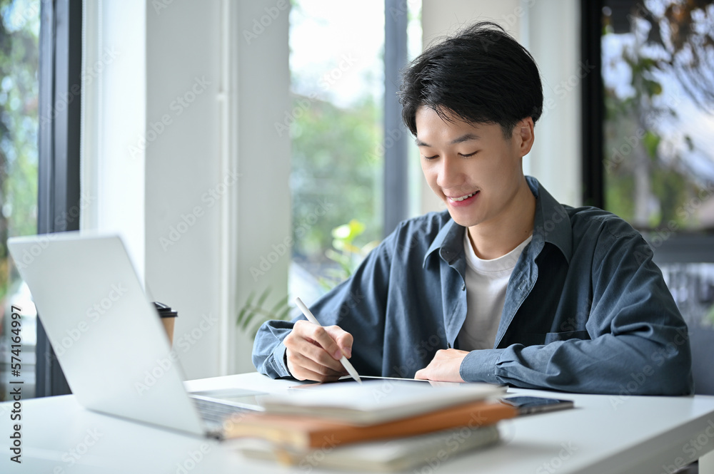 Happy and good-looking young Asian male college student designing his artwork on digital tablet Happy and good-looking young Asian male college student designing his artwork on digital tablet