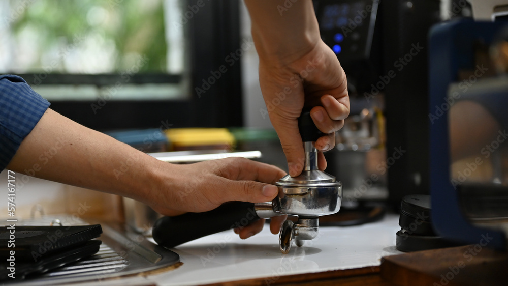 Professional male barista holding portafiler, using coffee tamper to press coffee powder