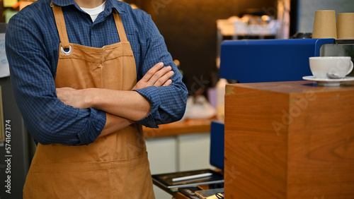 Successful Asian male barista in apron stands with arms crossed in the coffee shop.