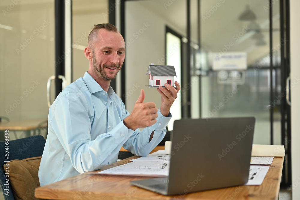 Confident man professional realtor holding house model offering new dwelling real estate to potential buyer via laptop