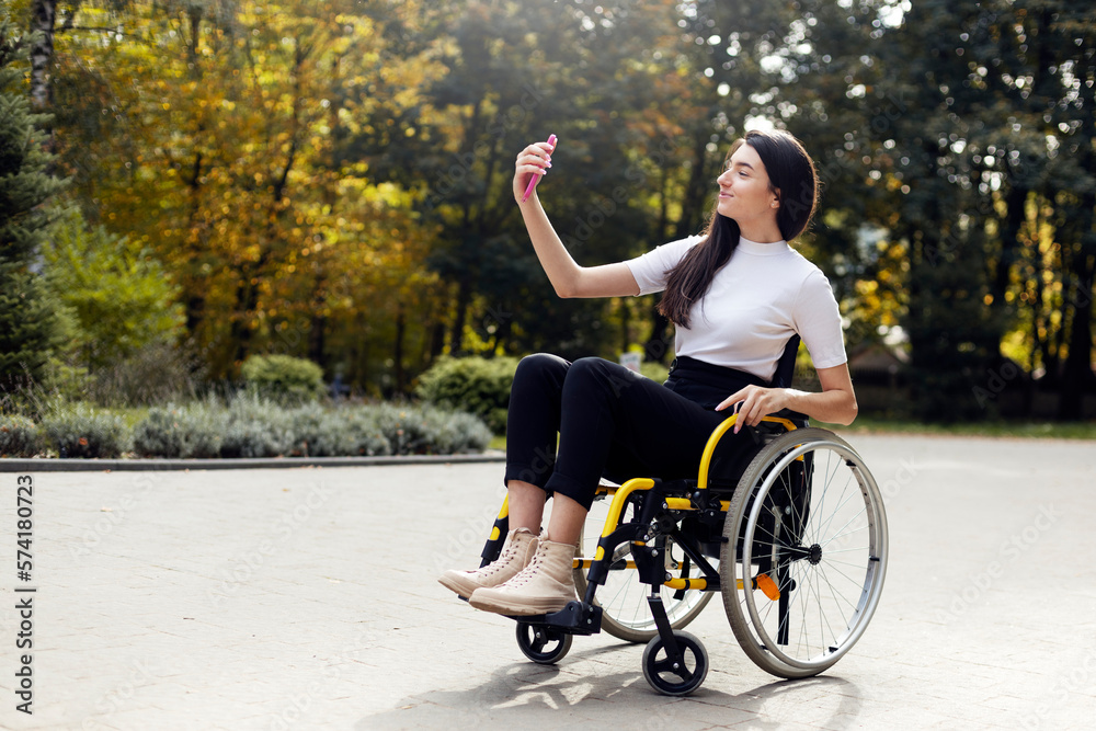 Attractive young woman sitting in a wheelchair with a phone in her hand. Beautiful girl smiling while taking a selfie on the phone outdoors. Attractive young woman sitting in a wheelchair with a phone in her hand. Beautiful girl smiling while taking a selfie on the phone outdoors.