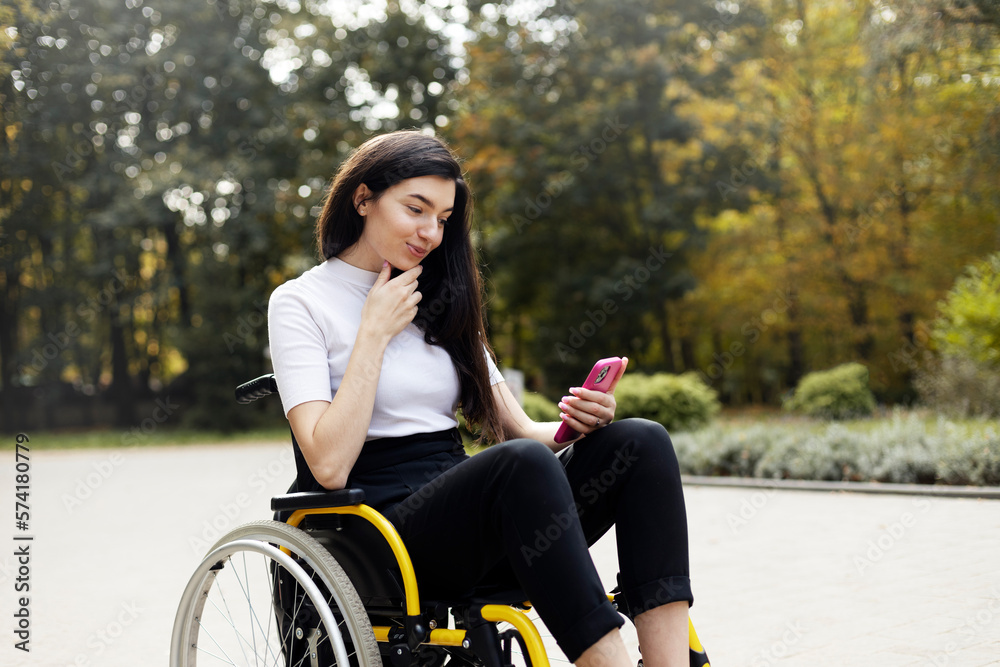 Cute, smiling brunette sitting in a wheelchair, looking at a smartphone. A beautiful, attractive woman - a disabled person on the street, in the park.