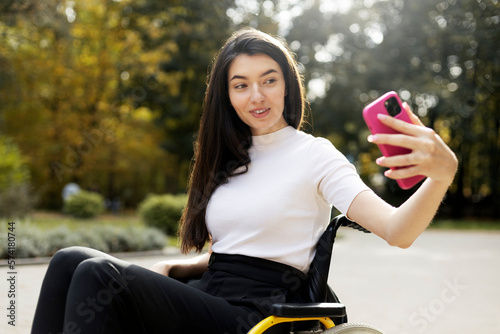 Charming, young woman holding a mibile phone in her hand, taking a selfie outdoors in the park. A beautiful girl sits in a wheelchair. photo