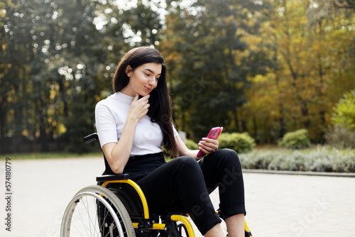Cute, smiling brunette sitting in a wheelchair, looking at a smartphone. A beautiful, attractive woman - a disabled person on the street, in the park. photo