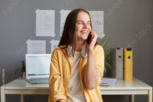 Photo of dreaming smiling woman with brown hair, fem ale wearing casual clothes and has headphones, talking via cell phone and looking away with happy expression. photo