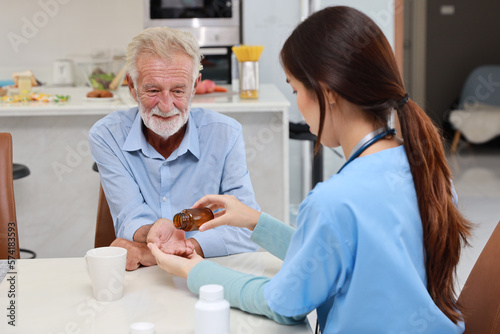 Happy caregiver sitting and helping senior caucasian man taking medicine after breakfast in living room at retirement house. Asian smiling nurse taking good help care and support elder patient at home