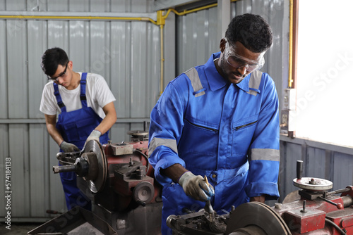 Multiethnic technicain mechanic or worker man in protective uniform using metal lathe machine operate polishing car disc brake at garage. Maintenance automotive and inspecting vehicle part concept photo