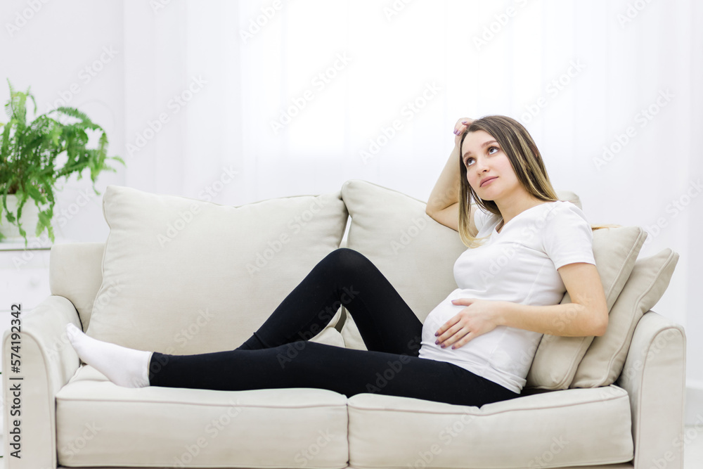Young pregnant woman thinking about something on white sofa. Young pregnant woman thinking about something on white sofa.