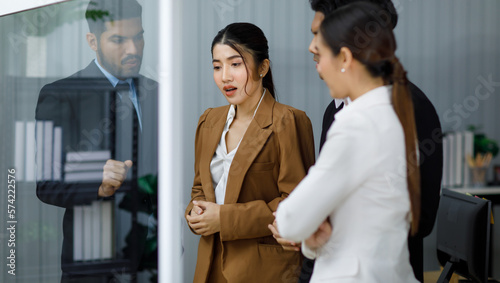Millennial Asian professional businesswoman employee staff officer in formal business suit standing crossed arms with male female colleagues listening to Indian businessman discussing on glass board