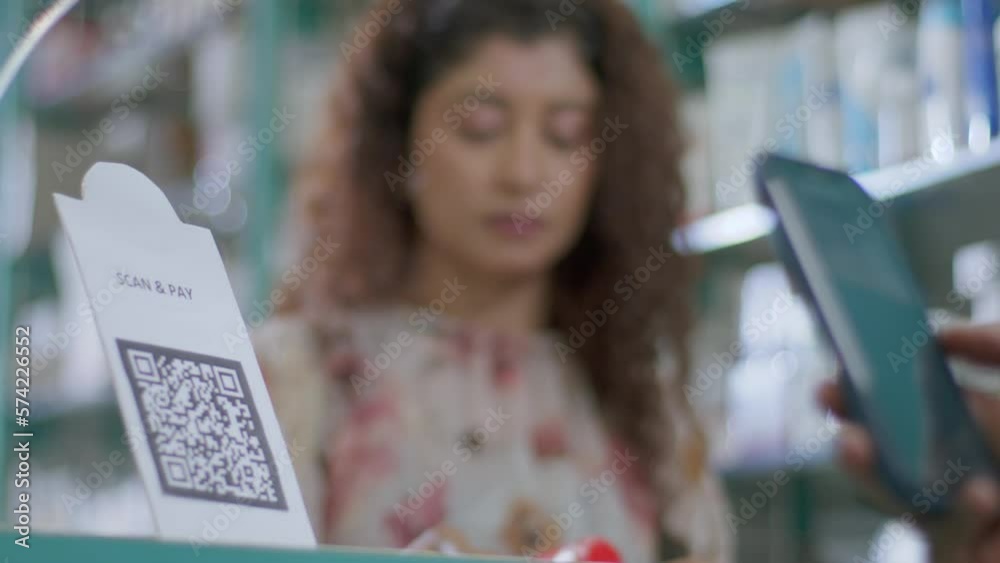 A close-up shot of a hand of a customer scanning a barcode to pay e-money to an electronic bank account or online UPI transfer in a general or grocery store with a smiling woman in the background