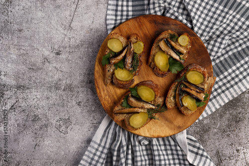 Sandwiches with sprats, herbs and pickles on a round cutting wooden board on a dark grey background. Top view, flat lay