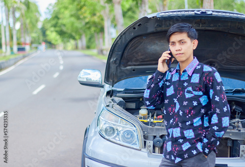 Confused handsome young Asian man wearing batik standing in the front car with a car engine breakdown and using mobile phone to call insurance. Accident emergency on the road concept