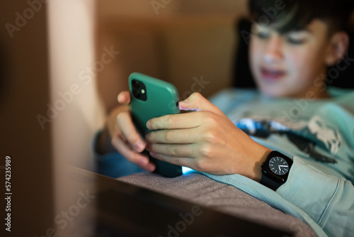 Young teen boy texting phone in front of a laptop on a bed at evening.