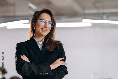 Portrait of smiling manager girl wearing glasses and black suit. Happy businesswoman standing with arms folded and looking away
