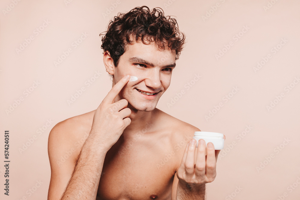 Close up portrait of young attractive smiling man with healthy skin, applies cream moisturizer on face for anti wrinkle or anti aging, cares of body, poses naked or shirtless, over beige background.