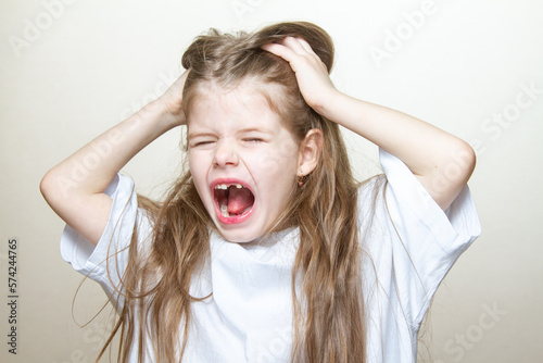 Portrait of a cute little blonde girl showing emotions, screaming and pulling her hair. A young girl screams in pain and holds her head.