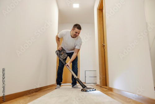 A happy neat man is using vacuum cleaner to vacuum carpet in hallway. © dusanpetkovic1