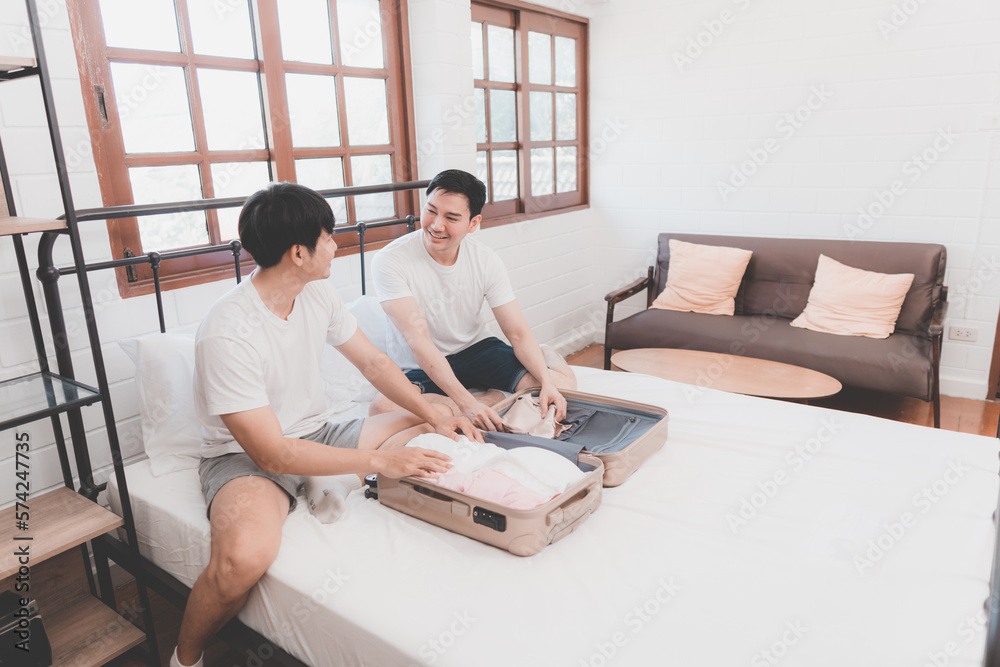 Happy gay couple packing suitcase for travel preparing for holiday travel. Diverse gay couple sit on bed packing luggage. laughing together planning booking hotel. Couple of gay tourists.