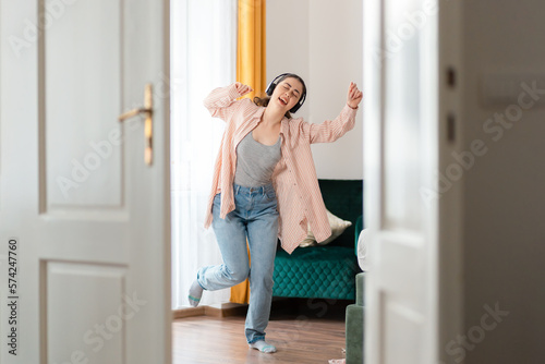 Free Download Happy young Caucasian happy woman wearing headphones dancing merrily in living room. Window in background. Leisure in home - GFXLoad