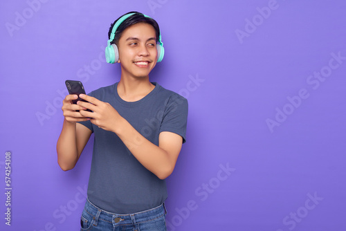 Happy young Asian man wearing casual clothes and headphones holding mobile phone, looking aside at copy space isolated over purple background © Bangun Stock Photo
