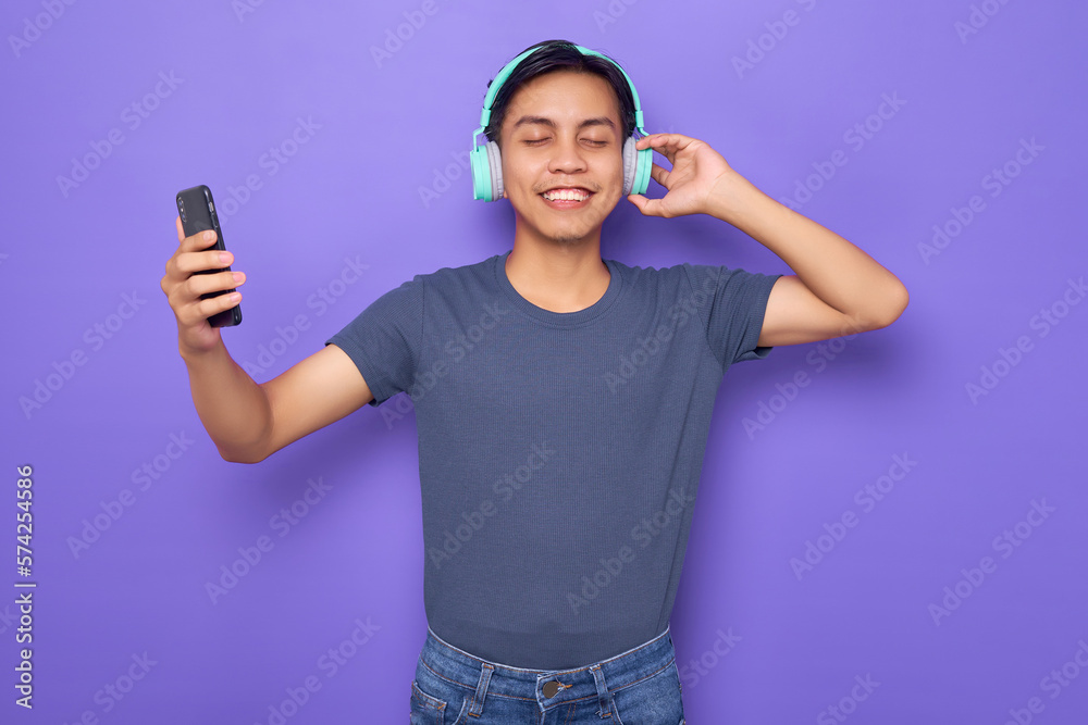 Young Asian man in a casual t-shirt wearing headphones for listening to music via mobile phone isolated over purple background