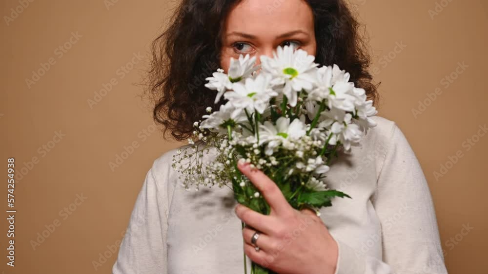 Close-up portrait of gorgeous brown-eyed middle-aged brunette, enjoying the smell of a spring bouquet of beautiful chrysanthemums and gypsophila flowers for Womens Day, on isolated beige background Close-up portrait of gorgeous brown-eyed middle-aged brunette, enjoying the smell of a spring bouquet of beautiful chrysanthemums and gypsophila flowers for Womens Day, on isolated beige background