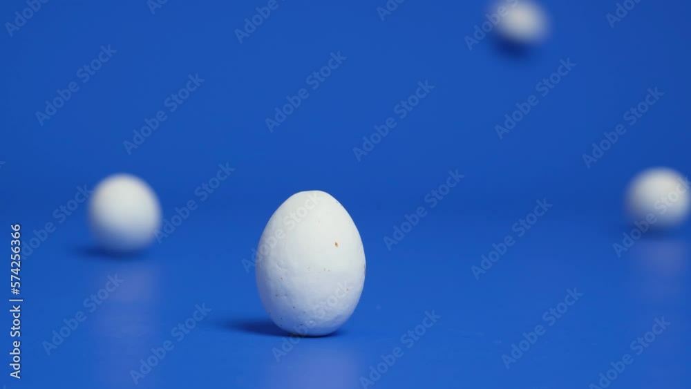 White chocolate Easter eggs roll across a blue background, knocking each other down. White chocolate Easter eggs roll across a blue background, knocking each other down.