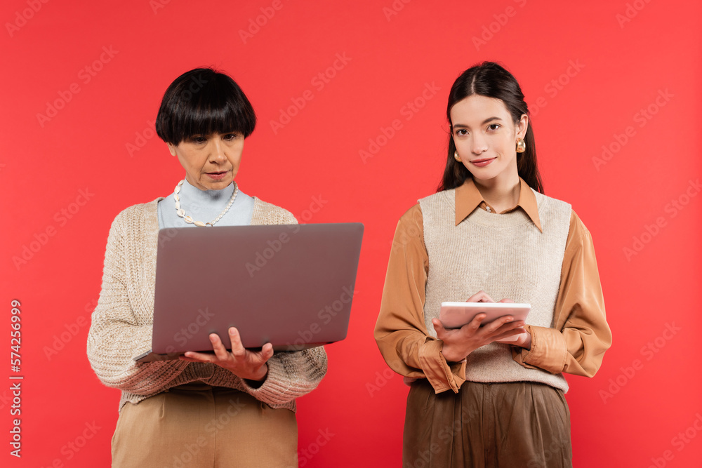 middle aged woman using laptop near young asian daughter holding digital tablet isolated on coral middle aged woman using laptop near young asian daughter holding digital tablet isolated on coral