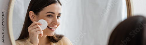 Canvas Print happy young woman cleansing face with cotton pad and looking at mirror in bathroom, banner