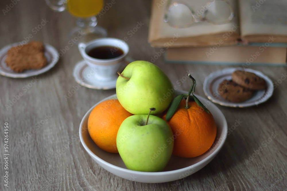 Cup of tea, plates with cookies, glass of orange juice, books, reading glasses, bowl of fruit and candles on the table. Selective focus.