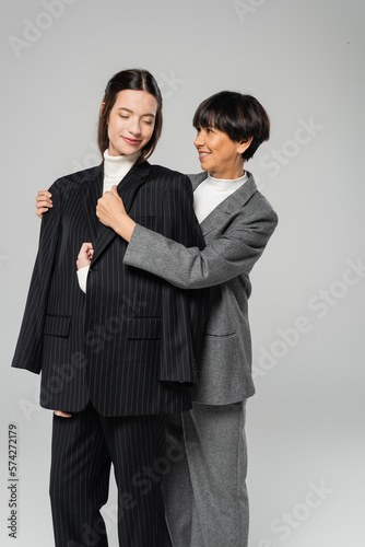 happy asian woman covering young brunette daughter with black blazer isolated on grey