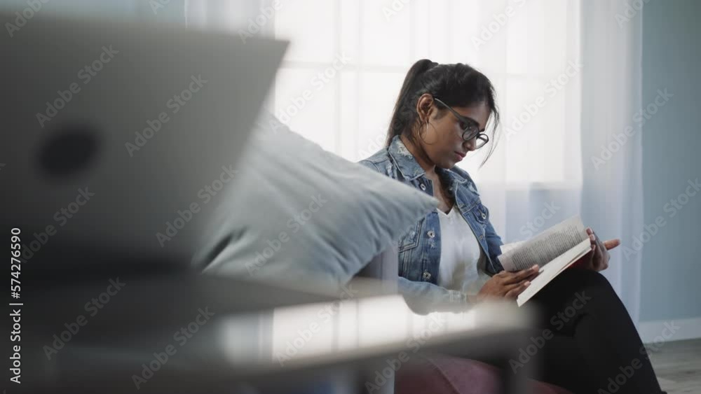 Attentive Indian student learns new information in textbook sitting in room against window. Brunette woman gets prepared for exams at home closeup